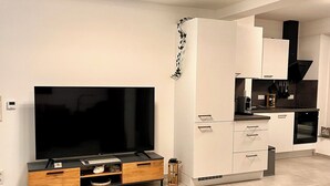 Een moderne woonkamer met een flatscreen televisie, een houten entertainmentkast en een keuken met witte kasten en zwarte apparatuur.