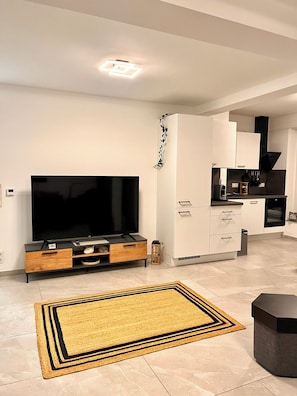 Uma sala de estar moderna com televisão de tela plana, um móvel de entretenimento de madeira e uma área de cozinha com armários brancos e eletrodomésticos pretos.