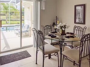 Uma área de refeições com mesa de vidro e cadeiras de metal, uma pequena mesa redonda com velas, e vista para uma piscina através de grandes portas de vidro.