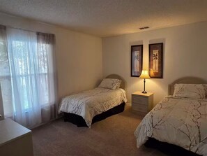 Quarto com duas camas, uma mesa de cabeceira, um abajur e uma janela com cortinas leves.