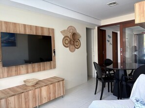 Ein modernes Wohnzimmer mit einem flachbildfernseher an einer Holzvertäfelung, einem Essbereich mit einem Glastisch und schwarzen Stühlen sowie einem dekorativen Wandkunstwerk aus Gewebe.