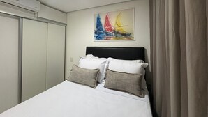 A hotel room with a bed, a painting of sailboats, a window with curtains, and an air conditioning unit. - Flamboyant Bessa Flat  (João Pessoa)