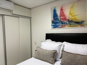 A bedroom with a bed, pillows, a painting of sailboats, and an air conditioning unit. - Flamboyant Bessa Flat  (João Pessoa)