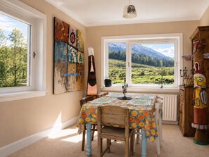 A dining area with a table set for four, a view of a mountainous landscape through large windows, and a rustic wooden cabinet with a carved figure. - Cragmhor Bed & Breakfast (Balquhidder)