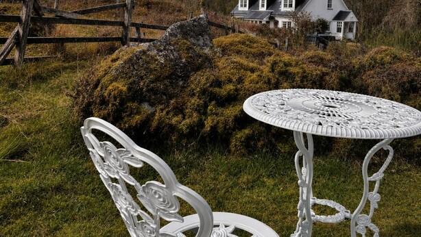 Conjunto de mesa e cadeira de metal branco em área gramada, com uma casa branca ao fundo.