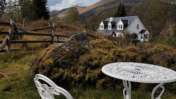 Une table et une chaise en métal blanc sont placées dans un espace verdoyant, avec une maison blanche en arrière-plan.