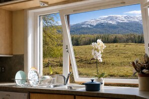 A kitchen with a view of a mountain landscape, a flower vase, and dishware on the counter. - CRAGMHOR (Balquhidder)