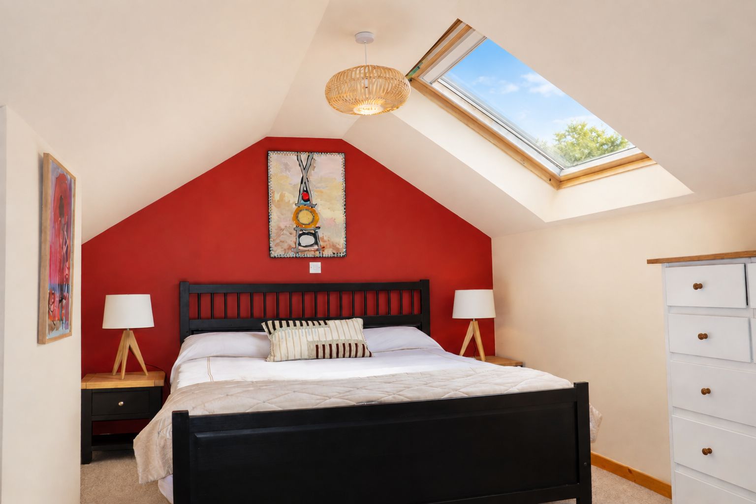 Une chambre avec un plafond en pente, un puits de lumière, un lit avec un cadre noir, deux tables de chevet avec des lampes et une commode à tiroirs.