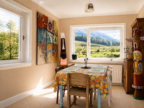 Un comedor con una mesa dispuesta para cuatro personas, vista a un paisaje montañoso a través de amplios ventanales y un aparador de madera rústica con objetos decorativos.