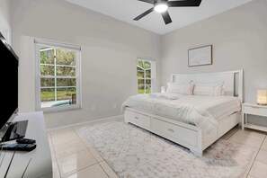 Um quarto com uma cama, um ventilador de teto, duas janelas e uma mesa de cabeceira.