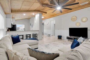 Uma sala de estar moderna com uma televisão de tela grande, um ventilador de teto e uma cozinha ao fundo.