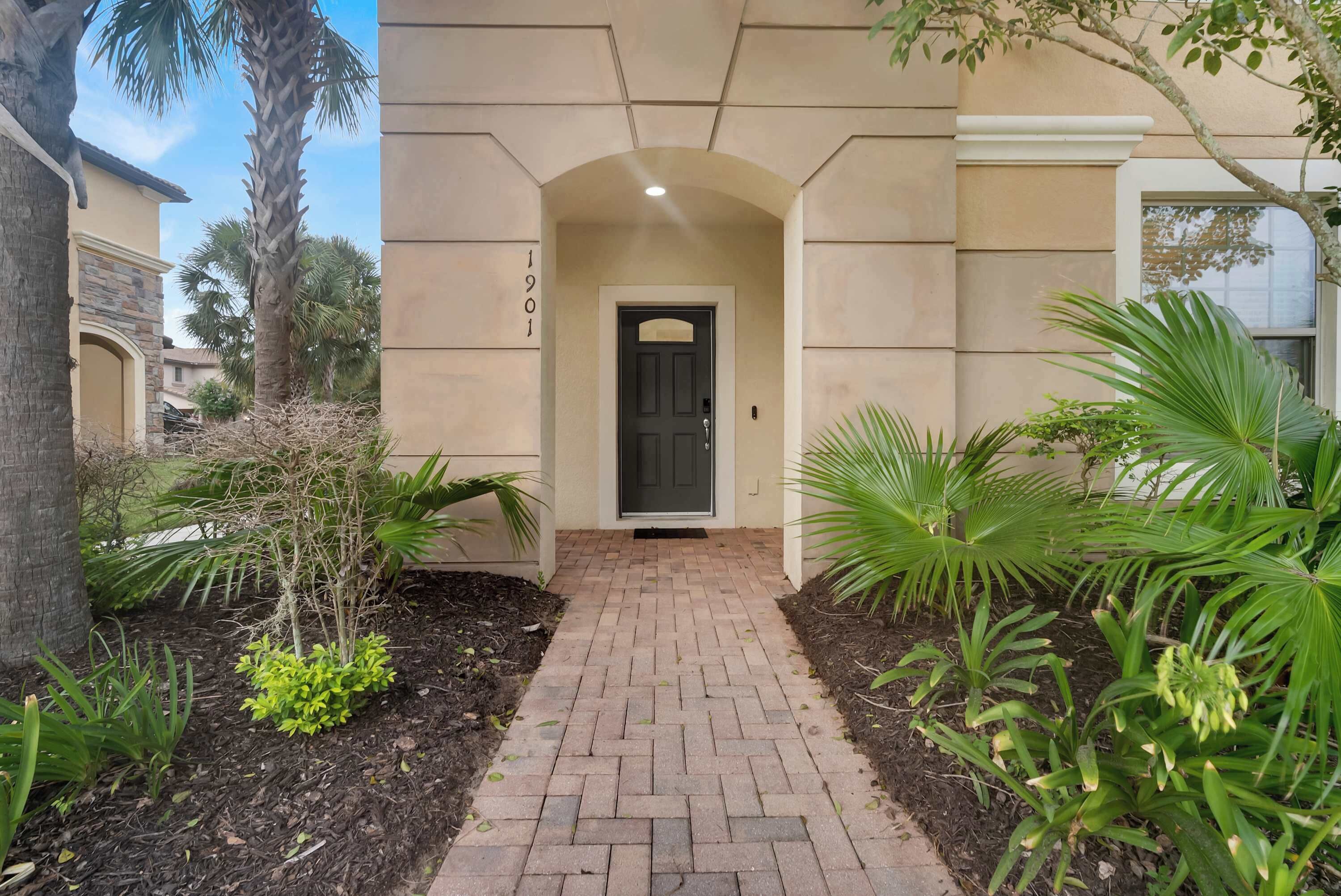 A paved entrance with a door, surrounded by landscaped greenery and a palm tree.