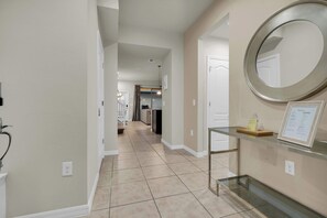 A hallway with a glass table displaying a welcome sign, a round mirror on the wall, and a door leading to a kitchen area. - 5BR Disney Escape with Pool and Game Room (Kissimmee)