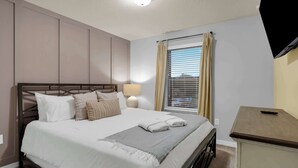 Un dormitorio con cama, lámpara de noche, ventana con persianas y televisor montado en la pared.