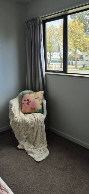 Une pièce d’angle avec une fenêtre, une chaise avec une couverture et un coussin, et un sol recouvert de moquette.