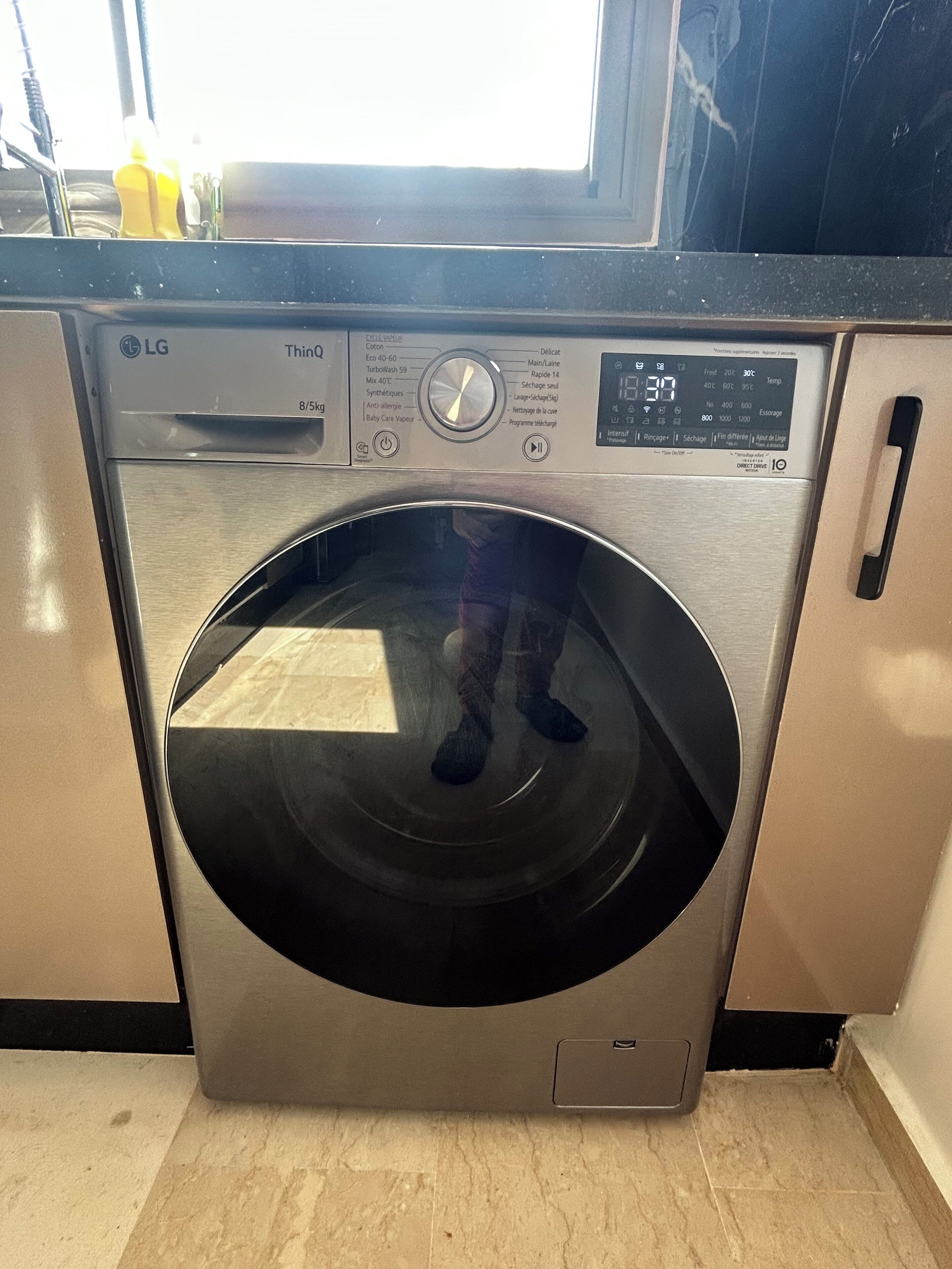 A front-loading LG washing machine with a control panel, located in a kitchen with a window above it.