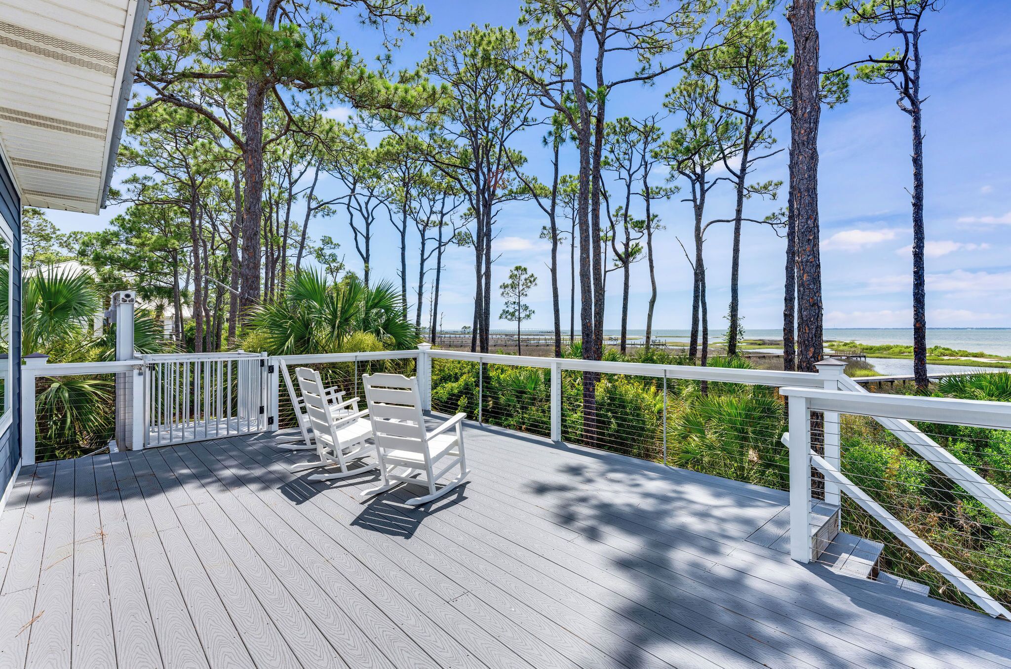 A wooden deck with white railings, two rocking chairs, and a view of the ocean and trees.