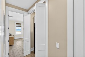 A hallway with white doors, a wooden floor, and a view of a beach through a window. - Gulf Watch! Across street from beach, private pool, elevator, and more! (St. George Island)