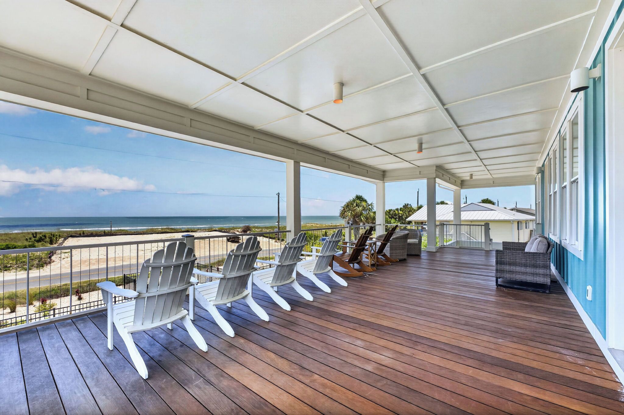 A covered outdoor deck with wooden flooring, white Adirondack chairs, and a view of the ocean.