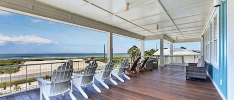 A covered outdoor deck with wooden flooring, white Adirondack chairs, and a view of the ocean.