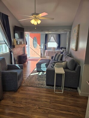 A living room with a sofa, a coffee table, and a ceiling fan. There is a door leading to another room. - Barking Bayside Bungalow (Lower Township)