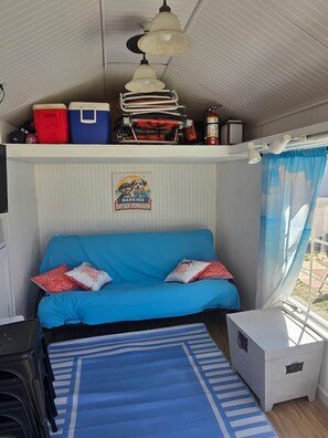 Een compacte kamer met een blauwe bank, een klein tafeltje, een plafondventilator en een raam met gordijnen.