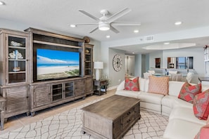 A living room with a large flat-screen TV, a ceiling fan, and a view of the kitchen and dining area. - CBC 2-6D - 2BR/2.5BA Gulf-view condo in Clearwater Beach, on Sand Key. (Clearwater Beach)