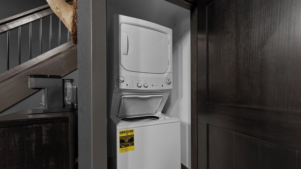 A laundry room with a stacked washer and dryer, a wooden staircase, and a caution sign on the dryer. - Lodge Cabin w/ Hot Tub | 4 Ensuite Kings (Hollister)