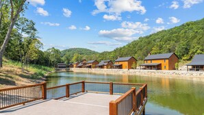 A wooden deck with a stone border, a calm river, and several wooden cabins with sloped roofs. - Lodge Cabin w/ Hot Tub | 4 Ensuite Kings (Hollister)