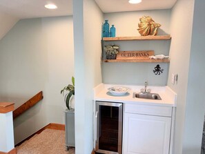 A compact kitchen with a built-in oven, a sink, and a small countertop. There are wooden shelves above the sink with decorative items and a sign that reads 'OUTER BANKS.' - OBXcitement: Oceanfront Condo, Water Views, Community Pool and Tennis Courts (Nags Head)