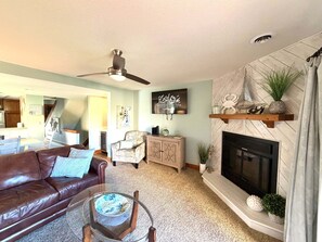A living room with a fireplace, a ceiling fan, a television, and a dining area. - OBXcitement: Oceanfront Condo, Water Views, Community Pool and Tennis Courts (Nags Head)