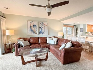 A living room with a brown leather sectional sofa, a glass coffee table, and two wall-mounted paintings of birds. - OBXcitement: Oceanfront Condo, Water Views, Community Pool and Tennis Courts (Nags Head)