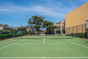 Cancha de tenis a l'aire lliure amb xarxa, envoltada per una tanca i adjacent a un edifici.