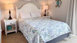 A bedroom with a bed, two bedside tables with lamps, a wall clock, a painting, and a curtain. - OBXcitement: Oceanfront Condo, Water Views, Community Pool and Tennis Courts (Nags Head)