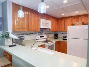 A kitchen with wooden cabinets, a microwave, a stove, a sink, and a refrigerator. - OBXcitement: Oceanfront Condo, Water Views, Community Pool and Tennis Courts (Nags Head)