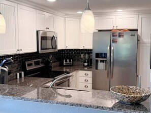 A modern kitchen with white cabinetry, a stainless steel refrigerator, a built-in oven, a microwave, and a granite countertop. - Carolina Treat: Oceanfront Condo, Quay Community Amenities, Quick walk to beach (Nags Head)