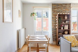 Ein Essbereich mit Tisch und Stühlen, einem Heizkörper, einem Fenster mit Vorhängen, einem Bücherregal und einem Sofa mit Kissen.