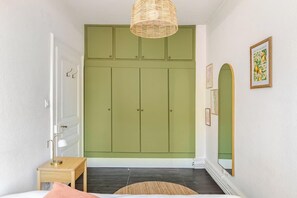 Une chambre avec une armoire verte, une table de chevet avec une lampe, un miroir et un tableau encadré accroché au mur.