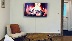 Una sala de estar con un televisor de pantalla plana montado en la pared, un sofá con cojines, una mesa de centro de madera y una puerta que da a otra habitación.