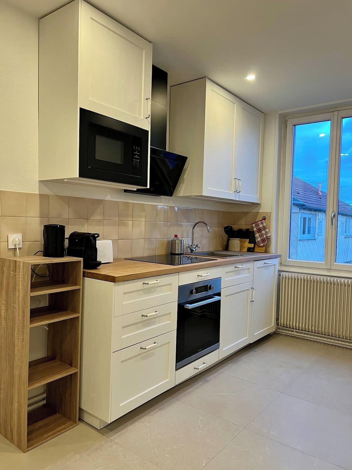 Une cuisine moderne avec des meubles blancs, un four à micro-ondes encastré, une machine à café et une fenêtre donnant sur une maison.