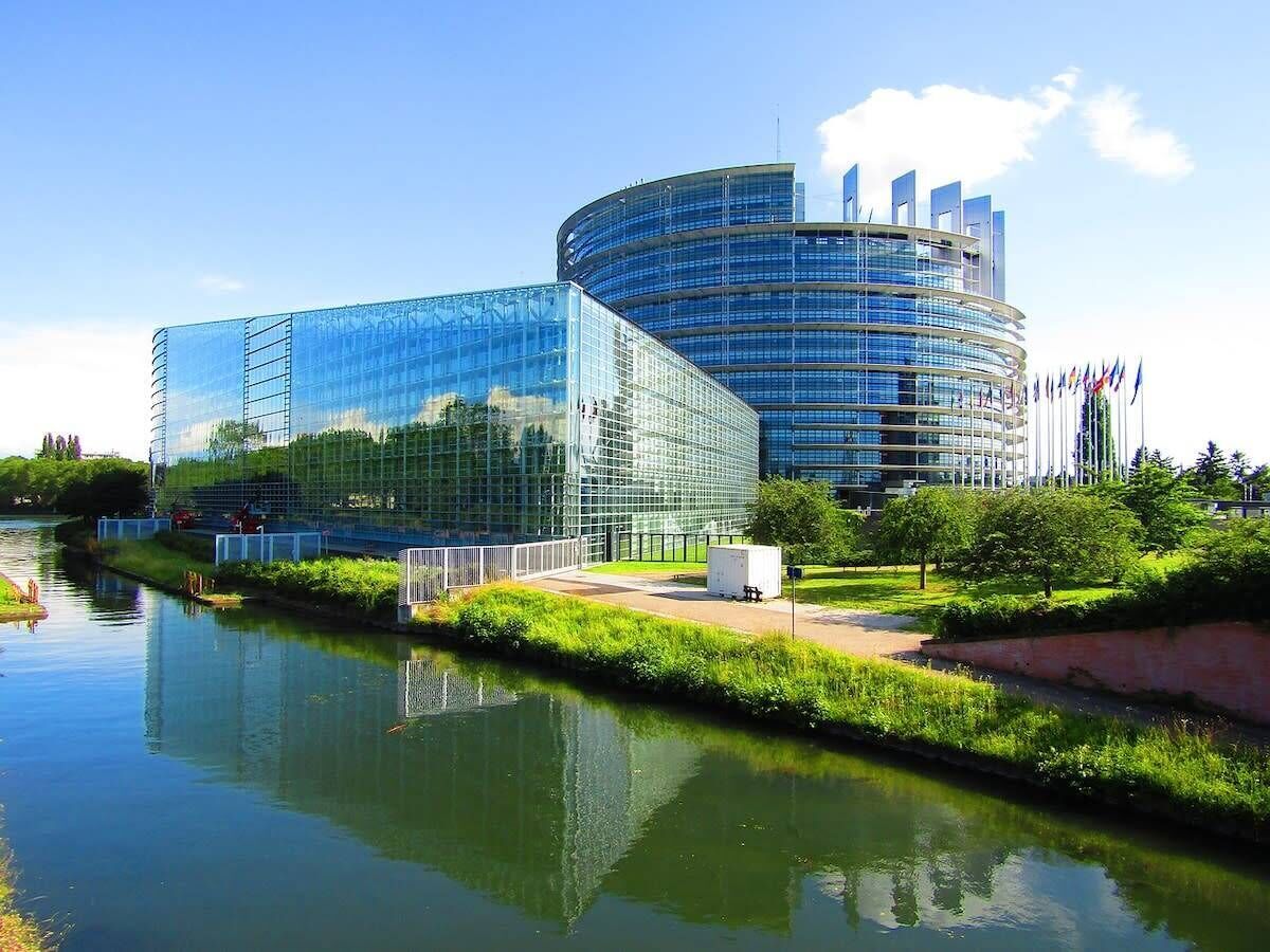 Un bâtiment moderne doté d’une façade en verre, d’un plan d’eau réfléchissant à l’avant et d’un mât sur lequel flottent plusieurs drapeaux.