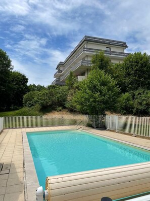 Una piscina con cerco blanco, rodeada de vegetación y un edificio de varios pisos al fondo.