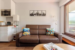 Ein modernes Wohnzimmer mit einem braunen Ledersofa, zwei bunten Kissen, einem Holztisch und einer Küchenzeile mit weißen Schränken und einem Edelstahlbackofen.