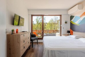 Ein Hotelzimmer mit einem Bett, einem an der Wand montierten Fernseher, einer Holzkommode, einem Sessel und einem Balkon mit Blick auf Bäume.
