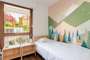 Une chambre à coucher avec un lit, une fenêtre donnant sur des immeubles et un mur orné d’une fresque représentant un paysage de montagne.