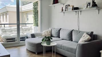 Een moderne woonkamer met een grijze bank, een klein wit bijzettafeltje en een potplant. Er staan witte wandplanken met boeken en decoratieve objecten.