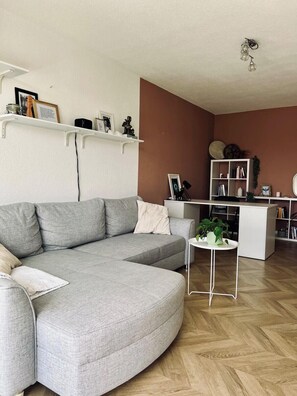 Ein modernes Wohnzimmer mit einem grauen Ecksofa, einem kleinen weißen Beistelltisch mit einer Pflanze und einer weißen Regaleinheit an einer braunen Wand.