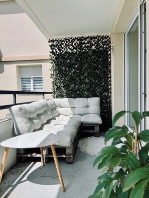 Ein Balkon mit einer weißen, gepolsterten Couch, einem kleinen runden Tisch und einem Holzstuhl. Eine grüne, blättrige Wand trennt den Balkon vom Gebäude.