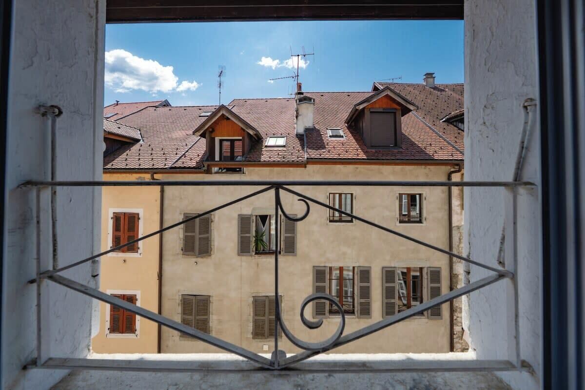 A view through a window with a metal railing, featuring a building with multiple windows and a tiled roof.