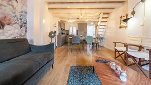 Una sala de estar con un sofá, un comedor con sillas, una cocina al fondo, vigas de madera en el techo y una pared con un mural floral.